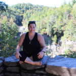Image of Leah Webb dressed in black yoga clothing, sitting on a rocky ledge in the mountains of North Carolina.