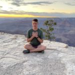 Garrett sits on the edge of the Grand Canyon with his eyes closed and hands are heart center